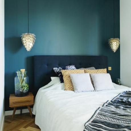 Stiilne magamistoa interjöör, millel on väike puidust öölaud, aed purgis, valge voodipesu, värvid padjad ja tekk. Ruum siniste seinte ja pruuni puitparketiga. Disainlamp.